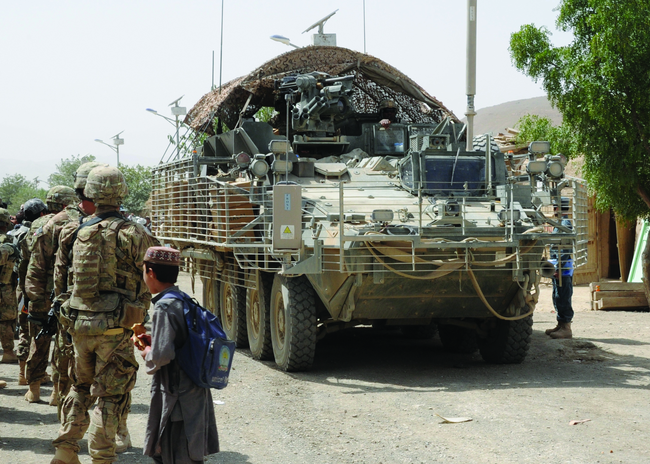 The rear view of a Stryker armored vehicle with camouflage netting, on patrol in a village street with soldiers and a local child nearby.